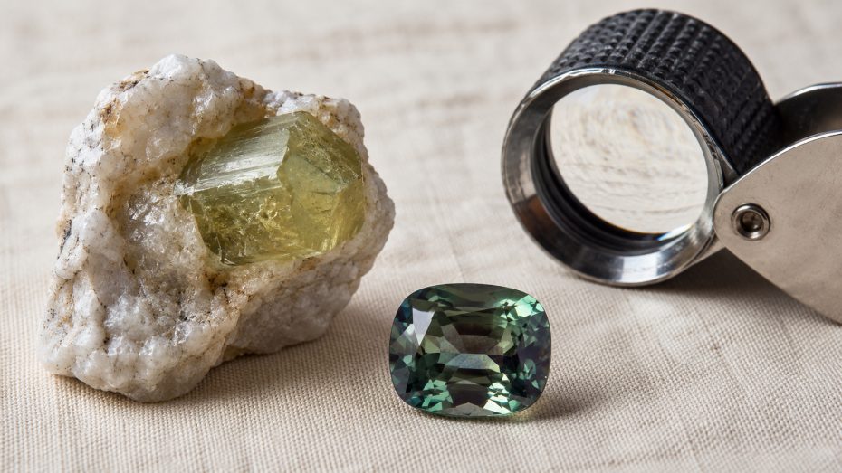 Roher Chrysoberyll-Kristall in hellem Muttergestein neben einem geschliffenen gr&uuml;nen Alexandrit und einer Juwelierslupe