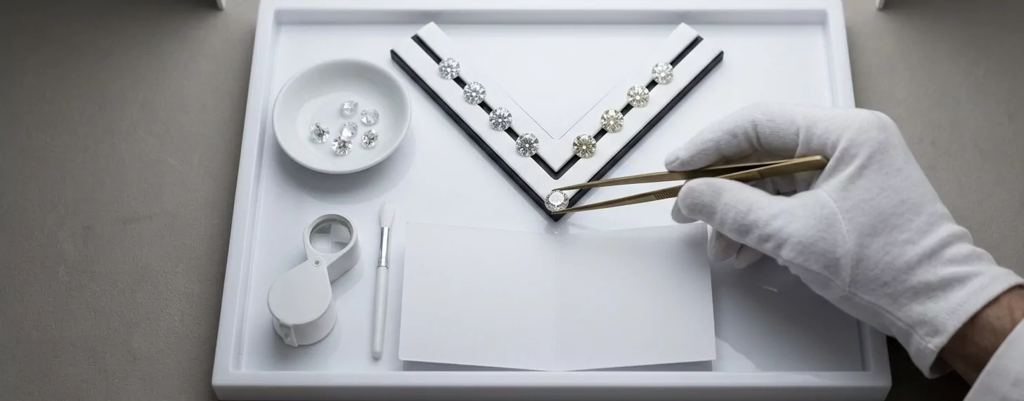 GIA-Bewertungs-Station von oben — weiß behandschuhte Gemmologen-Hand mit Messing-Pinzette über V-förmigem Master-Color-Set, kalibrierte 5500K-Tageslicht-Hood, Phillips-Auctioneers-Ästhetik.