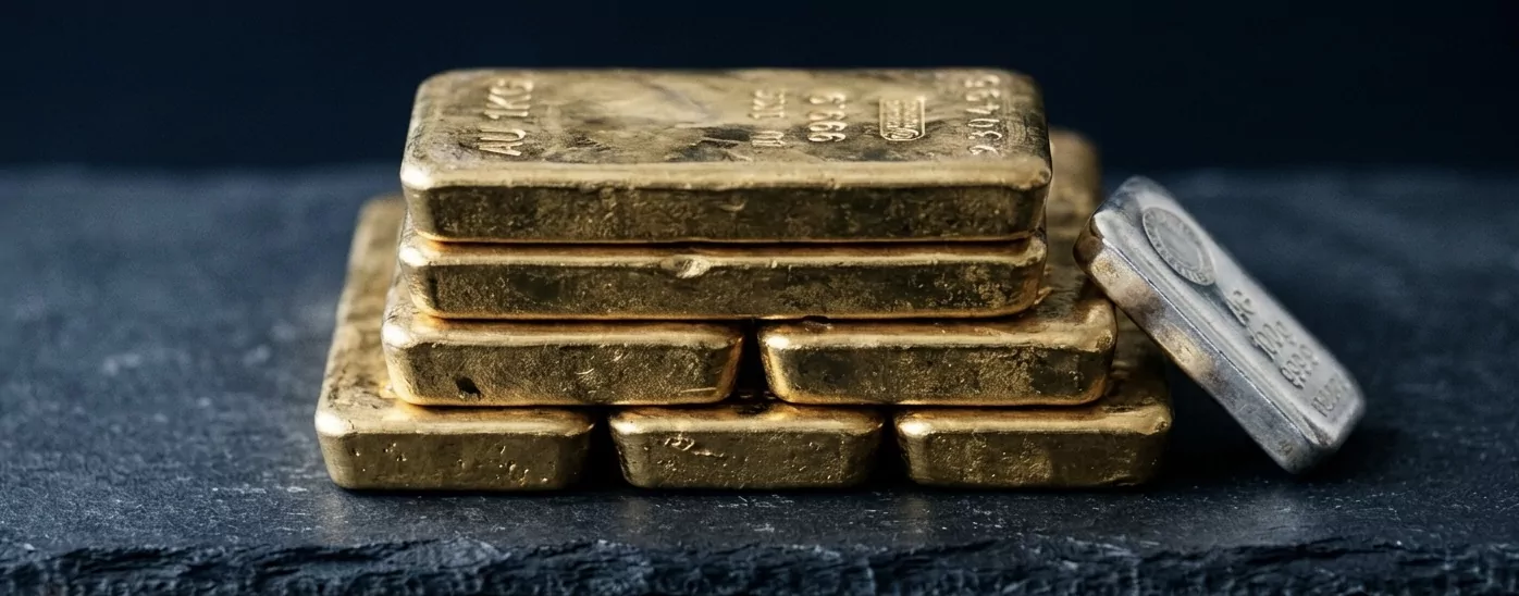 Stapel von sieben 1-Kilogramm LBMA-Good-Delivery-Goldbarren auf anthrazitfarbenem Schiefer, Assayer-Punze und Gewichtsstempel auf der Oberseite, einzelner 100g-Silberbarren leicht angelehnt zum Kontrast, Knight-Frank-Bloomberg-Markets-Editorial-Ästhetik.