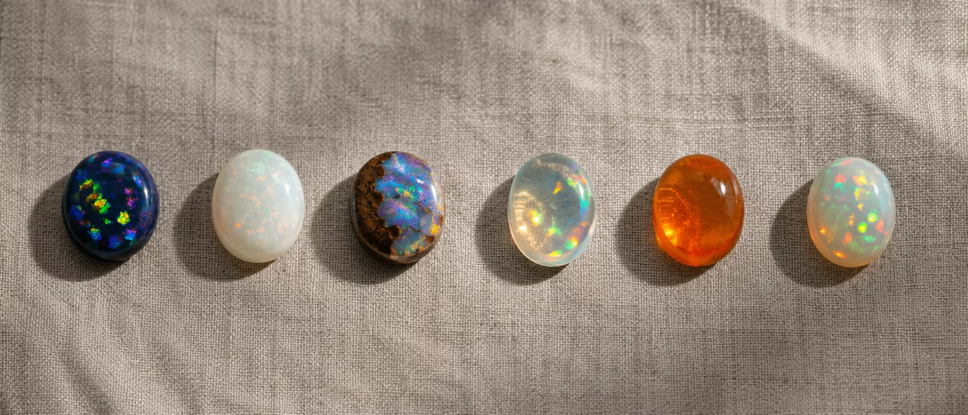 Vergleichsplatte sechs Opal-Familien auf sand-grauem Rohleinen — schwarzer Opal aus Lightning Ridge, weißer Opal aus Coober Pedy, Boulder Opal Queensland mit Eisenstein-Matrix, Kristall-Opal transparent, mexikanischer Feueropal orange, äthiopischer Welo-Opal translucent.