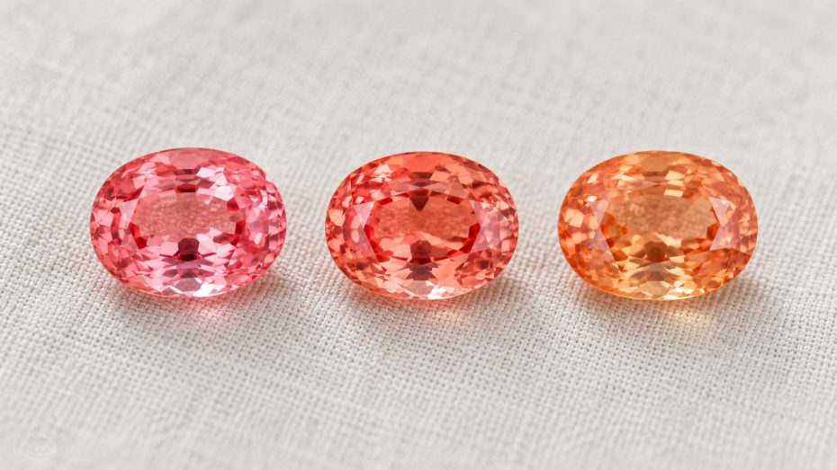 Drei Padparadscha-Saphire nebeneinander: einer mehr rosa, einer ausgewogen rosa-orange, einer mehr orange, auf cremefarbenem Leinen
