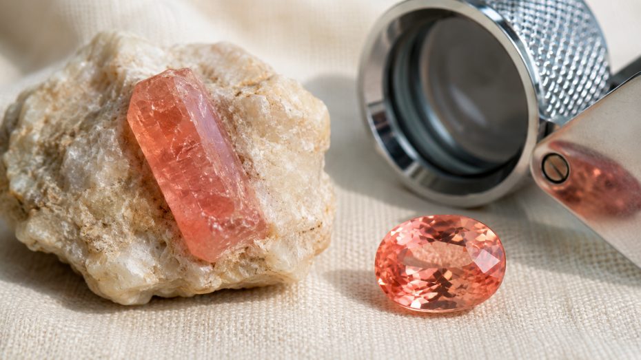 Roher rosa-oranger Korundkristall in hellem Muttergestein neben einem geschliffenen Padparadscha-Saphir und einer Juwelierslupe