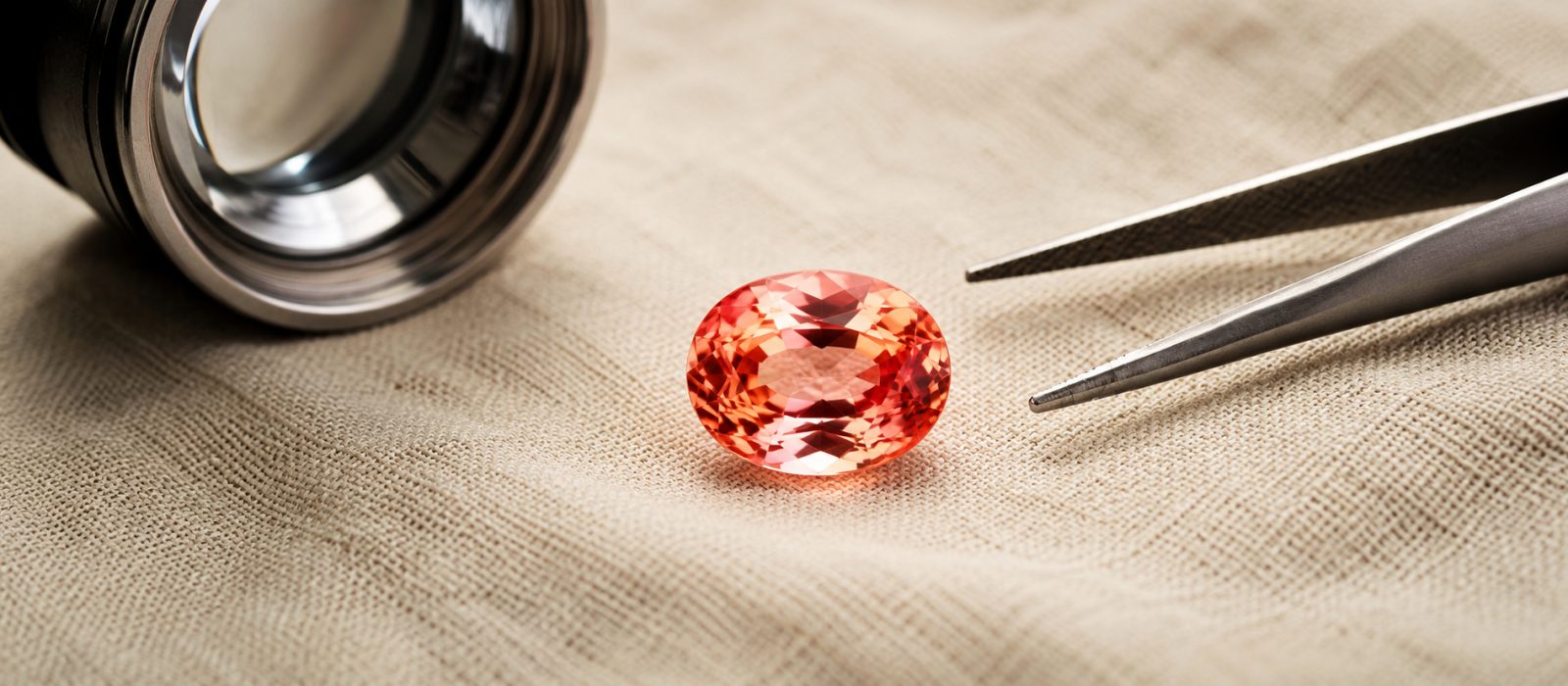 Loser Padparadscha-Saphir im Ovalschliff mit zartem Rosa-Orange, daneben Juwelierslupe und Steinpinzette auf cremefarbenem Leinen
