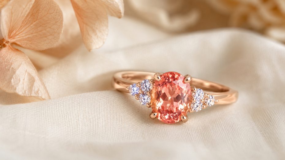 Ros&eacute;goldring mit ovalem Padparadscha-Saphir und seitlichen Brillanten auf cremefarbenem Stoff