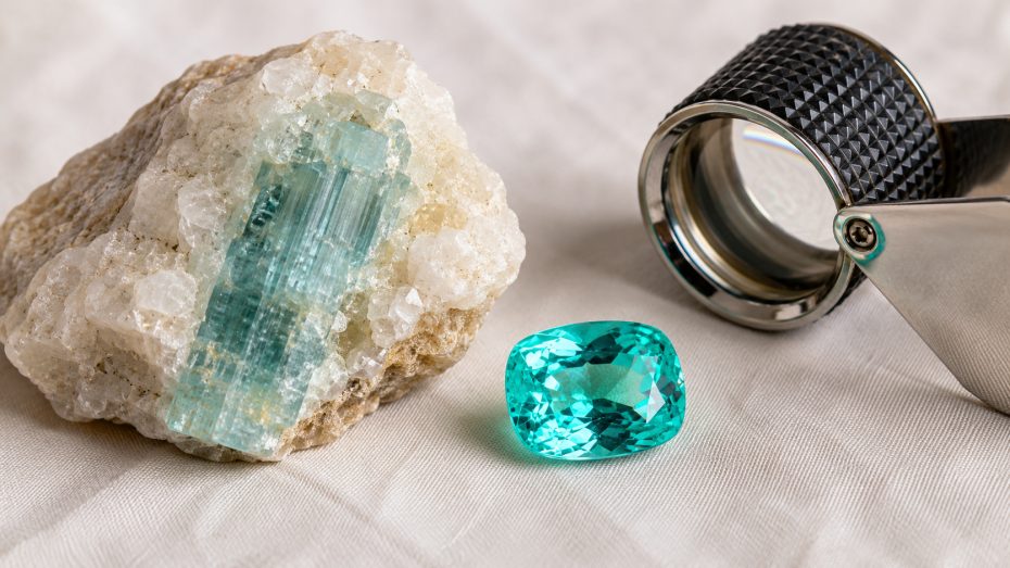 Roher Paraíba-Turmalinkristall in hellem Pegmatit-Muttergestein neben einem geschliffenen neonblauen Paraíba und einer Juwelierslupe