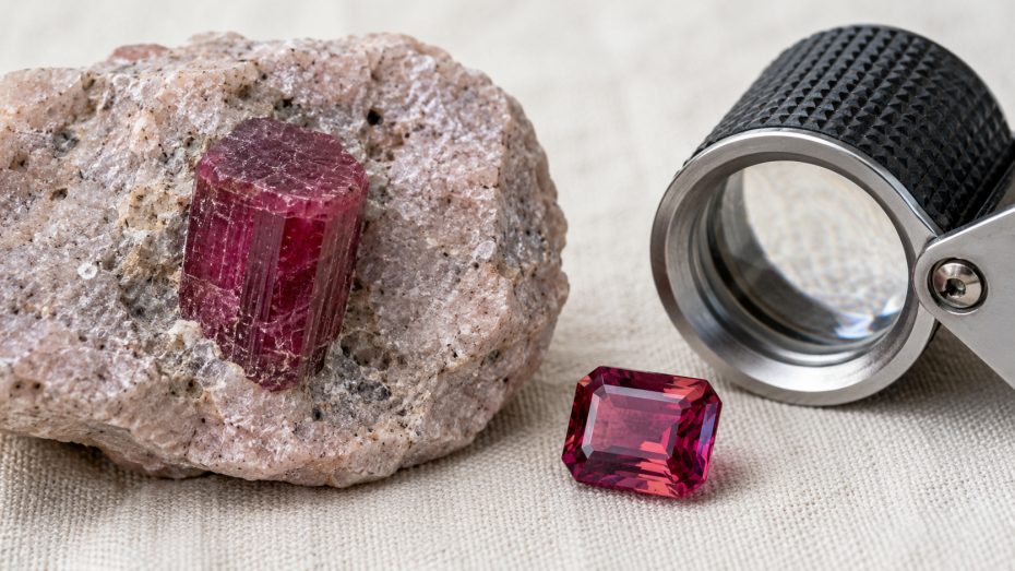 Roher sechseckiger Roter-Beryll-Kristall in hellem Rhyolith-Muttergestein neben einem geschliffenen himbeerroten Beryll und einer Juwelierslupe