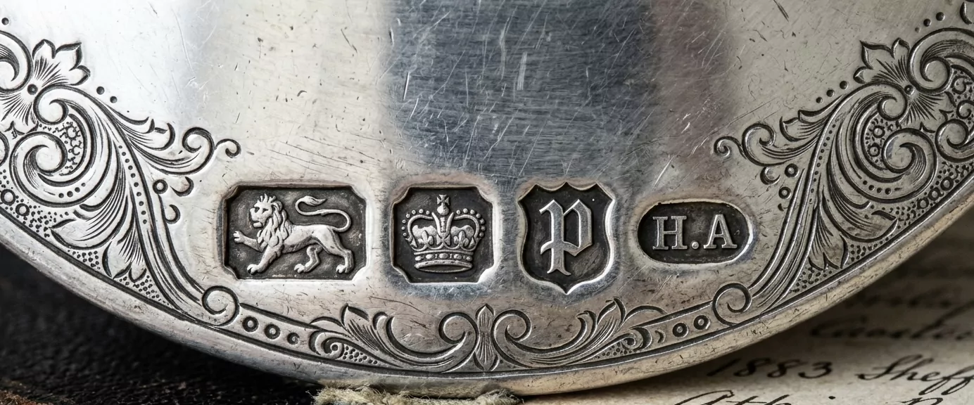 Britische Sheffield-Silber-Punzenreihe in extremer Makroaufnahme — Lion-Passant-Sterling-Mark, Sheffield-Krone-Stadtzeichen, gotischer Datumsbuchstabe P im Schild-Kartusche, ovale Meistermarke H.A, mit handgravierten dekorativen Bordüren.